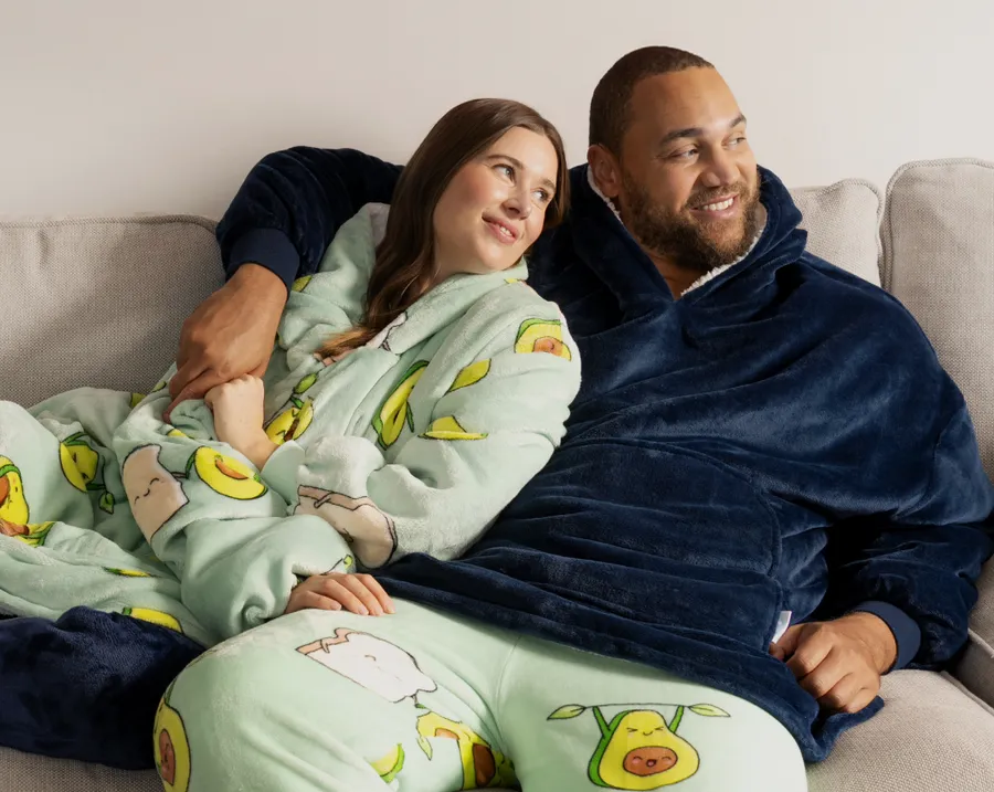 Two people cuddled up on a couch in Oodie™ Originals, hoods down and hands in the front pockets