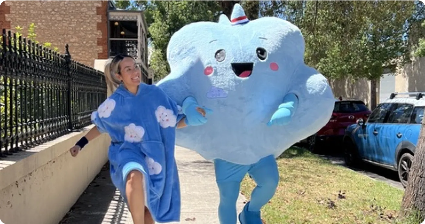 A person in a blue cloud Oodie walking down a street with a giant cloud mascot